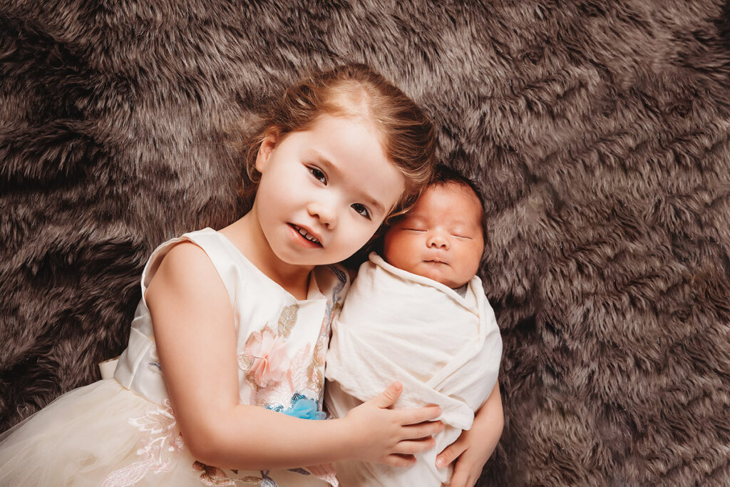 big sister wearing white floral dress holding newborn baby brother laying on brown fur
