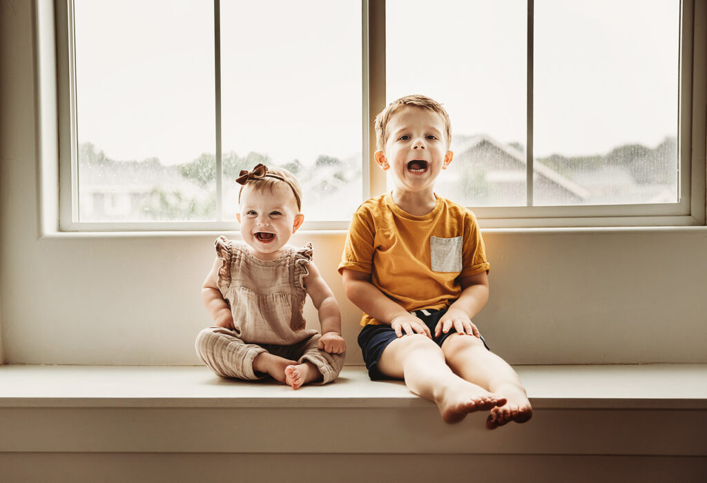 brother and sister grinning ear to ear sitting on a window seat