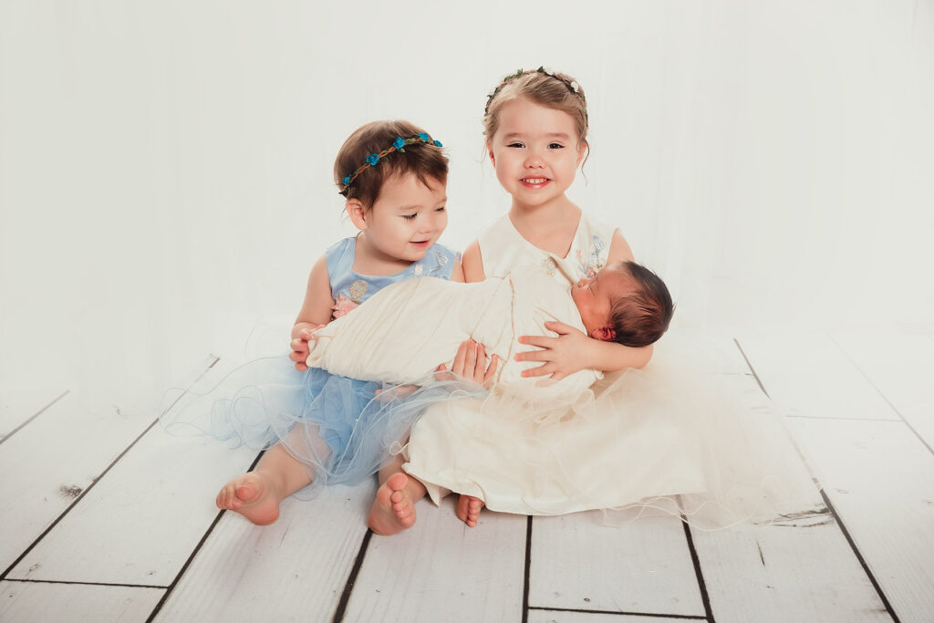 big sister wearing white dress sitting with little sister in blue holding newborn baby brother