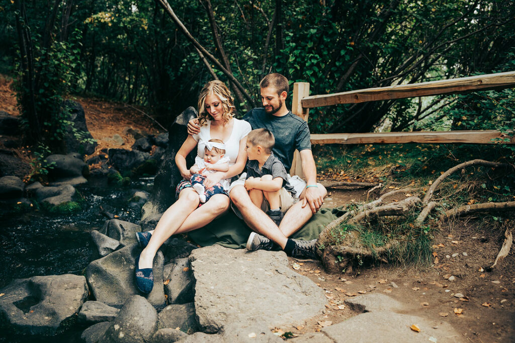 family of four sitting by the creek holding a baby and all looking at baby