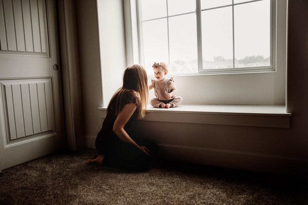 boise lifestyle photography little baby girl with ribbon in her hair sitting on window sill with mom kneeling next to her smiling