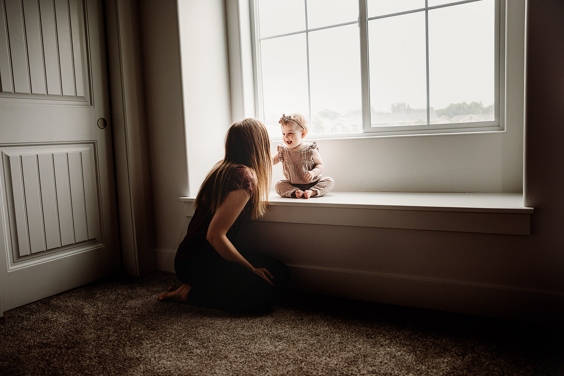 boise lifestyle photography little baby girl with ribbon in her hair sitting on window sill with mom kneeling next to her smiling