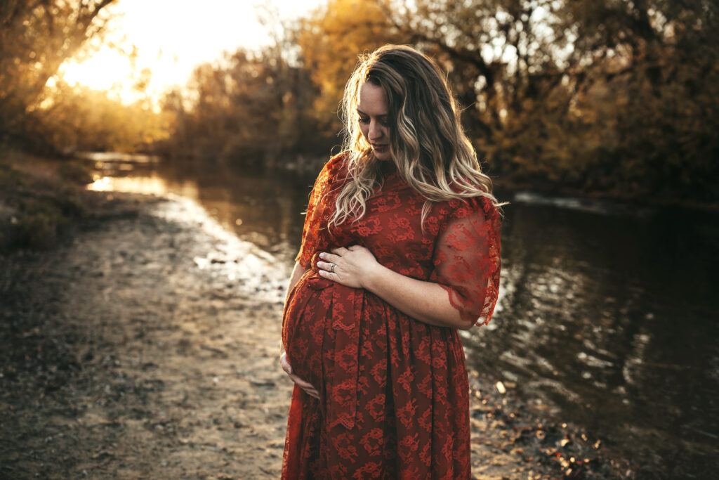 pregnant mama wearing lace maroon dress holding her belly and looking down standing on a river bank