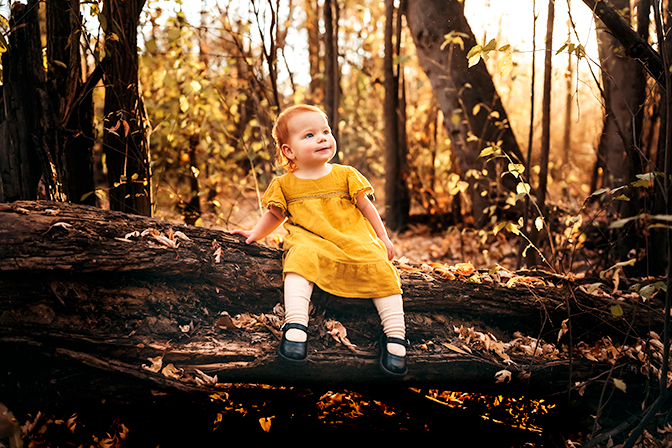 cute redhead baby girl wearing yellow dress sitting on a log smiling