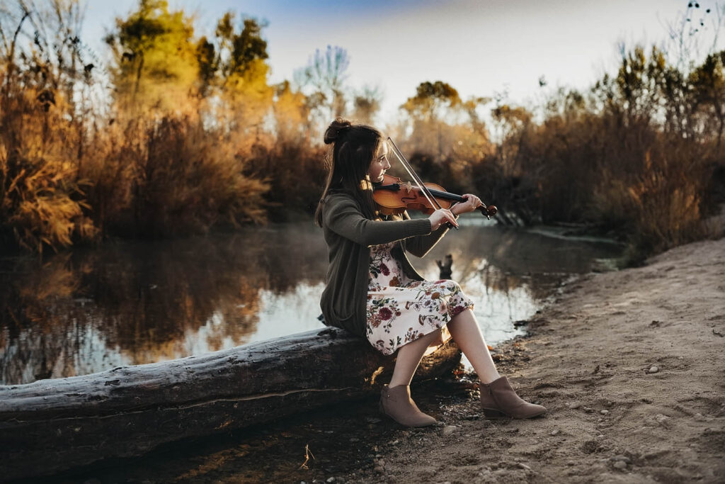teenage girl playing violin by a pond wearing a floral dress