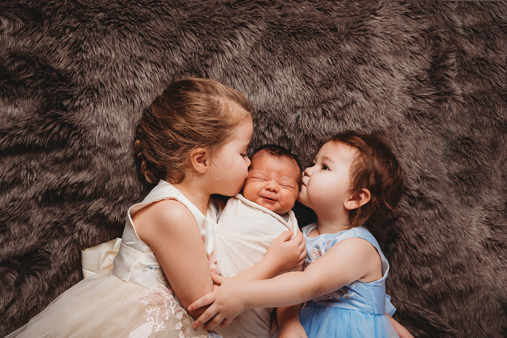 two sisters holding a newborn baby brother hugging him and kissing him on the head laying down
