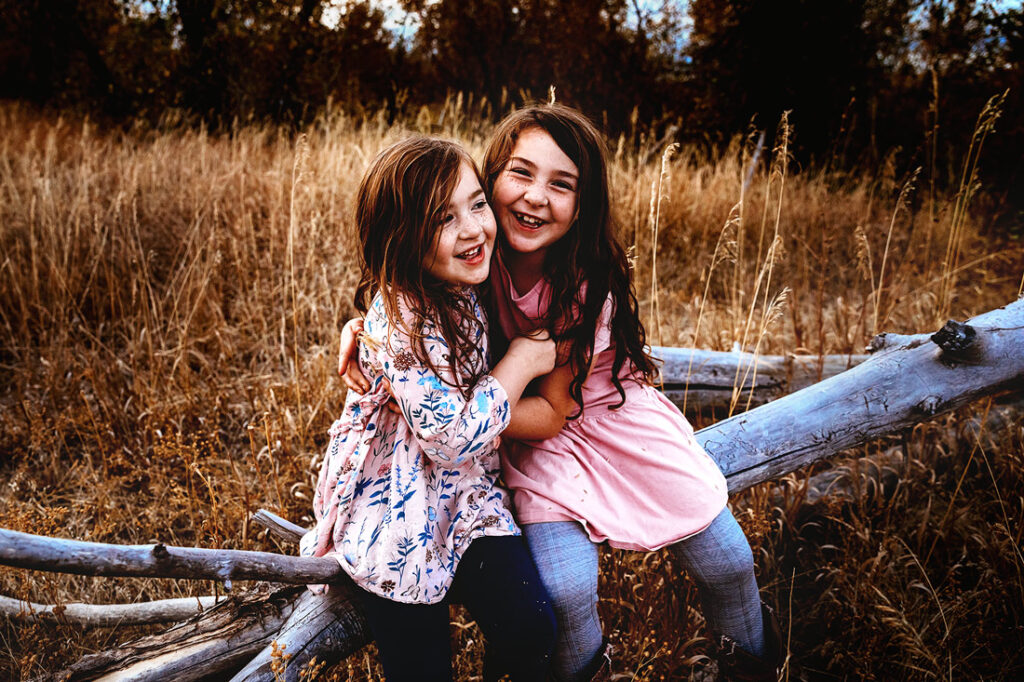 two sisters wearing pink tops hugging each other and giggling sitting on a log