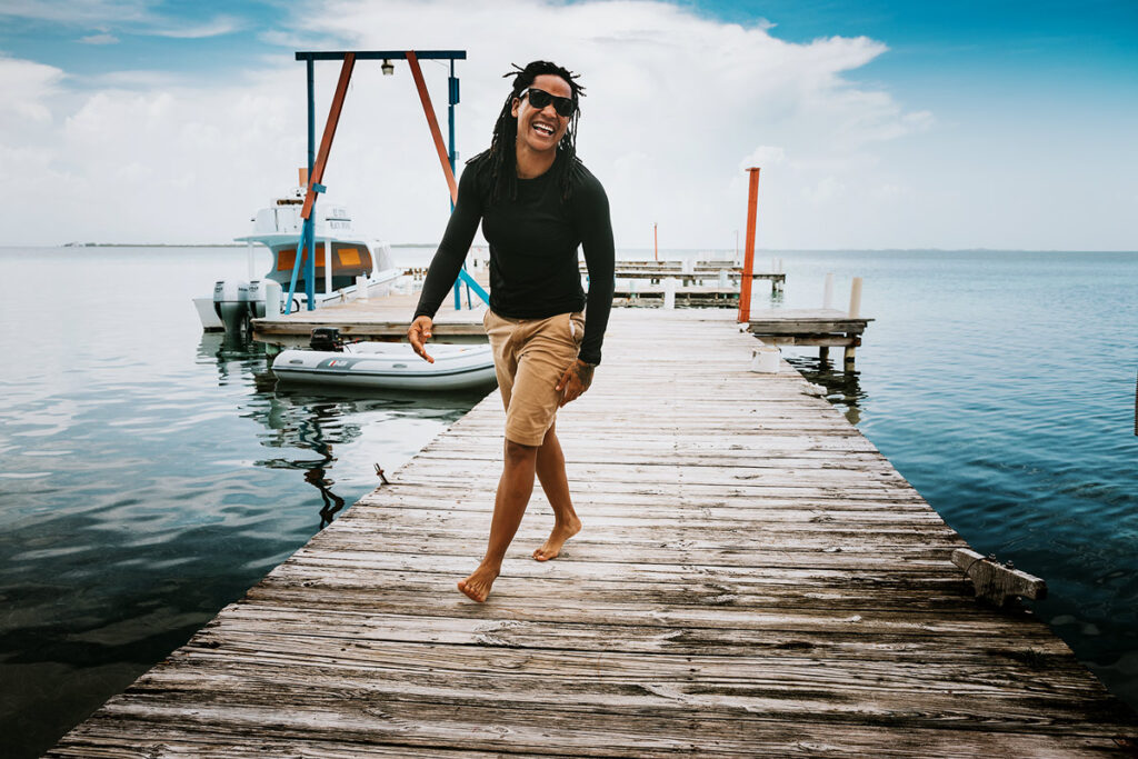 woman with dreads and sunglasses walking down a dock grinning with ocean and boats behind her